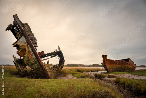 Decaying wooden shipwreck and rusting metal hull on marshland under overcast sky