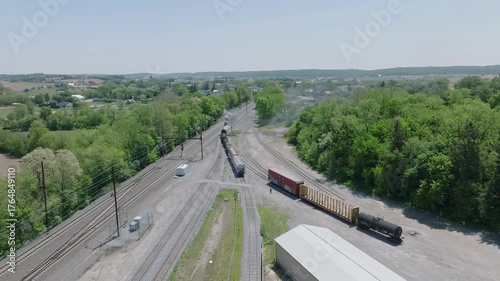 Steam Trains are moving through a bustling rail yard filled with containers and freight cars. The area is surrounded by trees and hills, creating a scenic backdrop under clear blue skies.