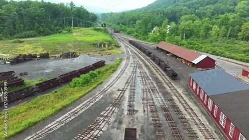 A wide view of an abandoned train yard reveals rusty train cars and old tracks. The serene landscape is filled with trees and distant mountains, creating a peaceful atmosphere.
