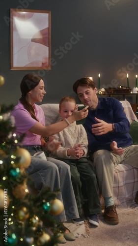 Vertical slowmo shot of smiling Caucasian family of three sitting together on cozy sofa and taking selfie beside beautifully decorated Christmas tree, enjoying festive holiday moments at home