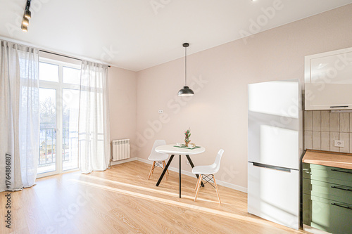 A bright, minimalist kitchen-dining area with light wood floors, a round table, white chairs, large windows with curtains, and modern appliances