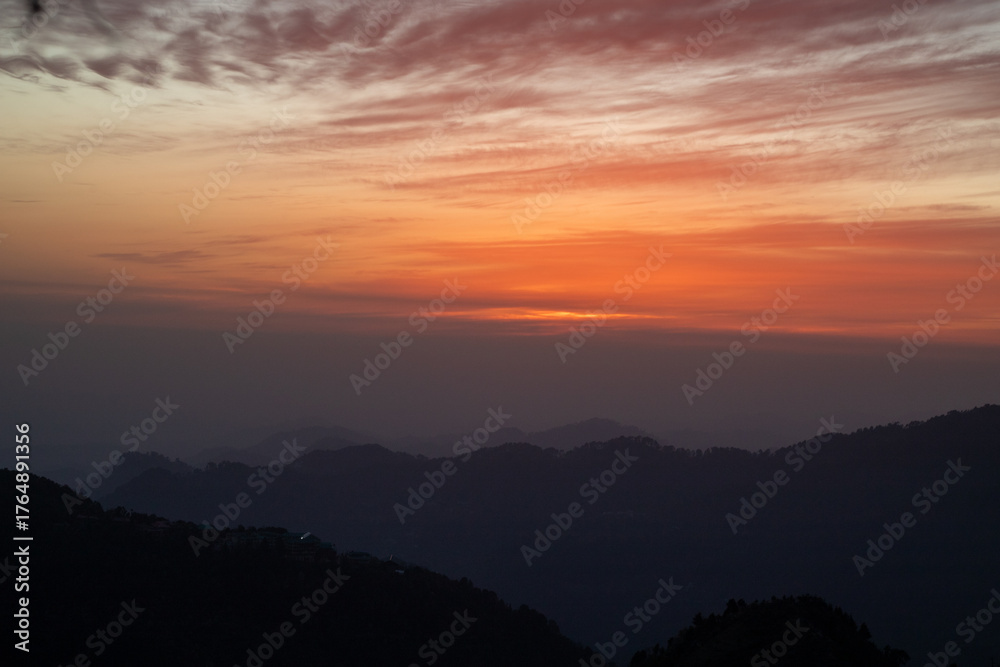 Fototapeta premium A breathtaking mountain sunset with layers of dark hills fading into the horizon under a dramatic orange and red sky. The soft glow of the setting sun and the textured clouds create a peaceful and ser