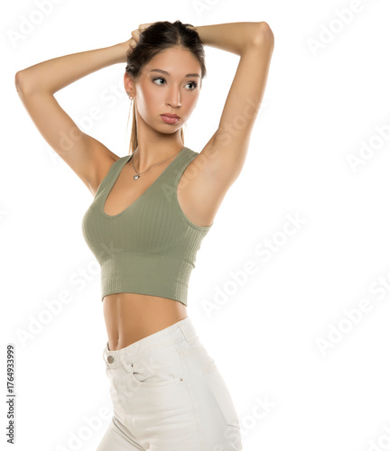 Studio shot of young woman with long brown hair in green top and white jeans making ponytail with arms raised on white background.