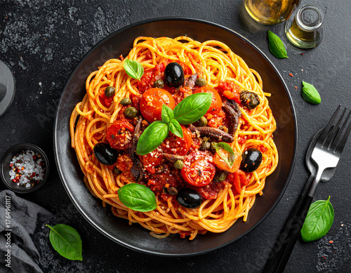 assic Italian Spaghetti alla Puttanesca with tomatoes, black olives, capers, and basil, served in a black bowl on a dark textured background with olive oil and a fork.