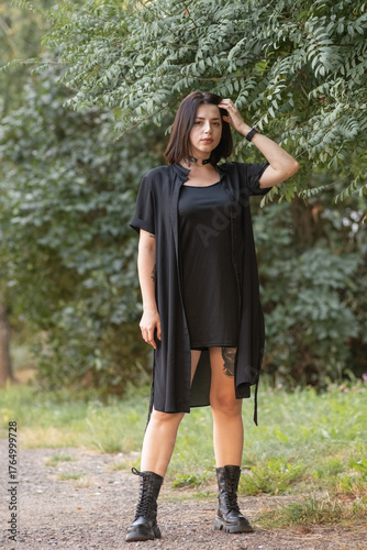 Confident woman in black attire stands on a forest path, holding straps of her outfit, surrounded by lush greenery, showcasing a bold fashion statement and natural beauty