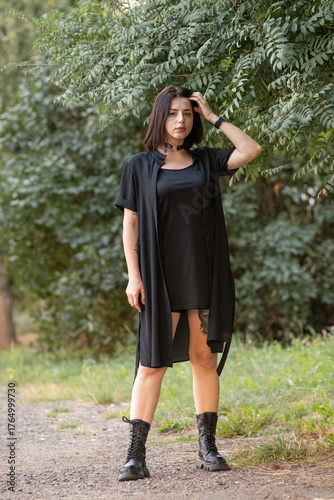 Confident woman in black attire stands on a forest path, holding straps of her outfit, surrounded by lush greenery, showcasing a bold fashion statement and natural beauty