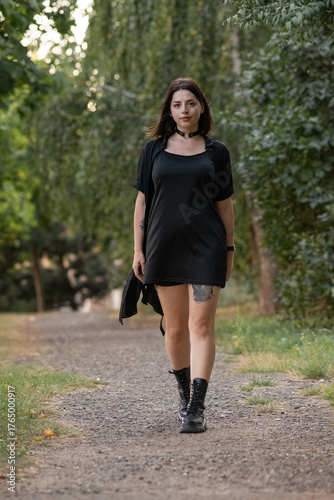 Female model in black clothing poses confidently on a forest trail, holding outfit straps, with vibrant greenery creating a serene and stylish atmosphere