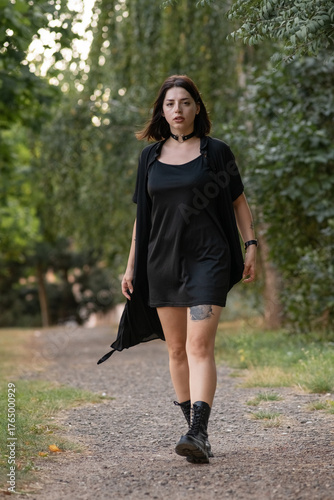 Female model in black clothing poses confidently on a forest trail, holding outfit straps, with vibrant greenery creating a serene and stylish atmosphere