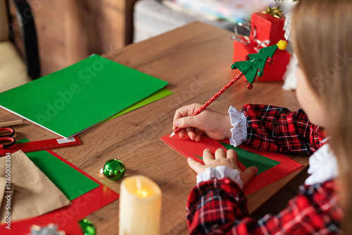 Child writing christmas card at festive table with decorations, holiday magic moment, festive crafting at home