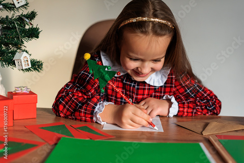 Young caucasian girl writing christmas cards in festive red dress by tree, Christmas wish letter to Santa Claus, Childhood creativity and festive spirit