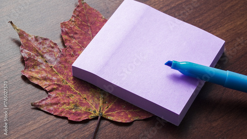 Purple sticky note and blue marker on autumn leaf resting on wooden table