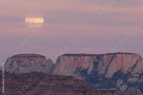 A full moon rises at sunset from behind the Alter of Sacrifice in Zion NP Utah USA, partly hidden by thin clouds which are stained pink from the last light of the day.