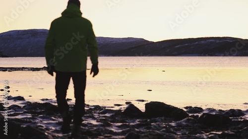 Man in winter clothing walks thoughtfully across ice crusts on the shore of the Atlantic Ocean during an icy winter in Northern Norway near Tromso