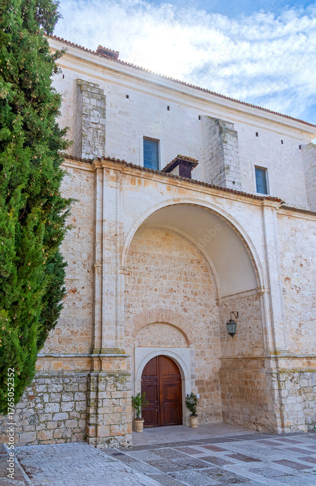 Naklejka premium The imposing white stone facade of the Church of Our Lady of the Assumption in Chinchón, Spain, featuring cypress trees, an arched entryway, and a stone cross