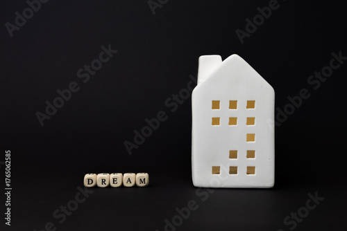 White ceramic house model with illuminated windows beside wooden blocks spelling dream, symbolizing aspirations and financial goals in real estate. copy space