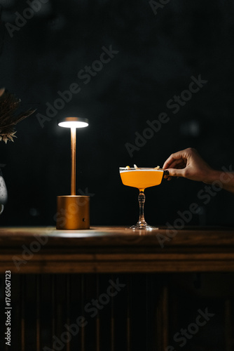 Stylish yellow cocktail in an elegant glass on a wooden bar with warm lighting and a golden lamp. Minimalist scene with a woman's hand reaching for the drink.

