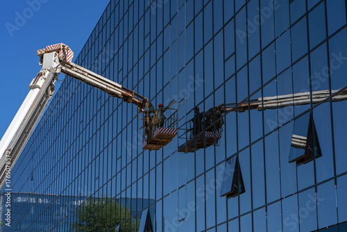 A construction crane with a cradle or lifting platform is reflected in the façade glazing of an office building. A construction worker in the crane bucket works on the façade glazing.