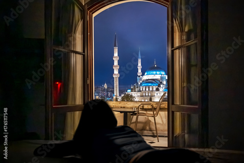 Photography A woman in silhouette lies on a bed and looks out a balcony window at the Galata Tower and the illuminated Yeni Mosque, also known as the New Mosque at night in Istanbul, Turkey