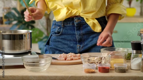 A woman in a yellow shirt and jeans is cooking in a kitchen with various ingredients and spices on the counter. Preparing lunch or dinner in a home kitchen.