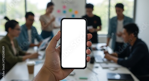 A person's hand holds a smartphone with a blank white screen in the foreground during a business team meeting in a blurred office.