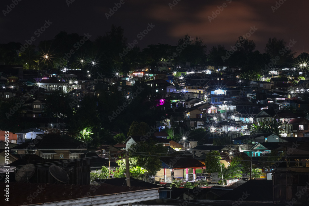 Fototapeta premium Night view of a dense, tiered residential neighborhood on a hill, illuminated by streetlights and house lights with beautiful starburst effects.