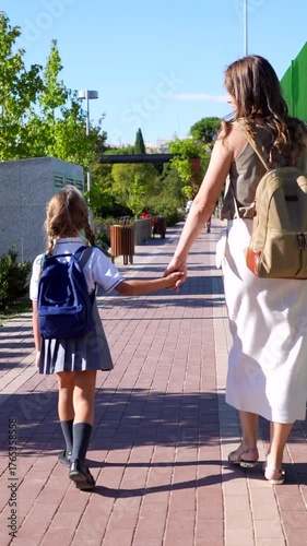 Back view of a mother taking her little daughter to school on the first day of class, both carrying backpacks and walking hand in hand along a sunlit city pavement on a beautiful day