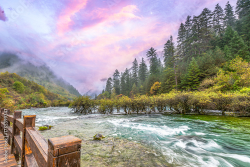 Misty forest morning along a wooden boardwalk beside a turquoise mountain river in Jiuzhaigou National Park, Sichuan, China. Tranquil autumn landscape under pastel pink sunrise skies.