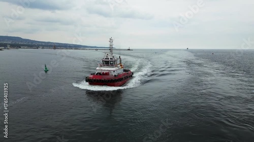Aerial view of a red tugboat sailing through the port channel toward open sea. Maritime navigation and harbor operations concept