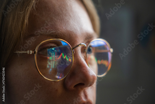 Фотография Close-up portrait of a focused young woman trader with glasses reflecting financial charts, long blond hair and fresh natural face, working at a desktop with screen glow in dim, shadowy lighting