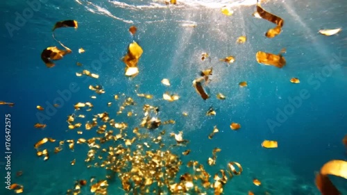 Underwater view of golden confetti and streamers falling through clear blue water with sun rays creating a festive and magical atmosphere.