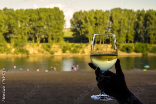 Frosted glass of cool white wine in woman hand, against blurred picturesque background of river beach with people. Close-up.