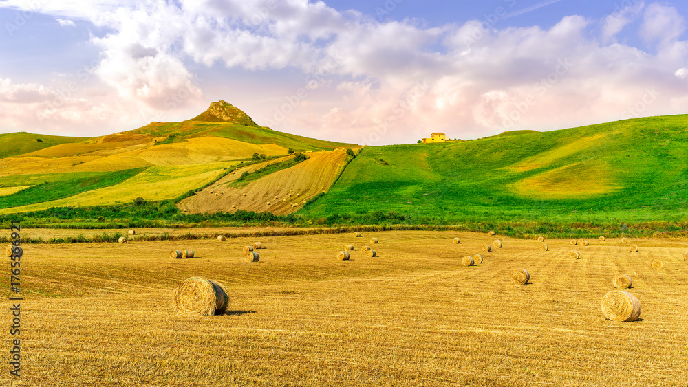 Obraz premium Scenic view at picturesque golden sunset valley in a green shiny field with hay stacks, bright cloudy sky , golden sun rays and beautiful hills on background, summer valley landscape