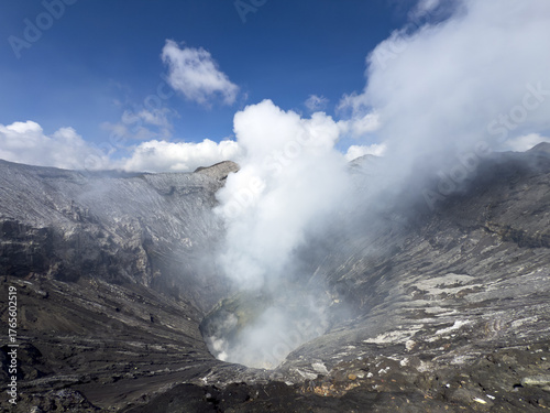 View of a smoking volcano crater with ashen slopes under a vibrant blue sky dotted with fluffy white clouds, Bromo, East Java, Indonesia.