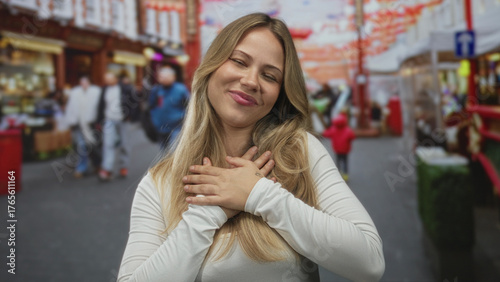 Free Download Woman blond smiling warmly placing hands on chest among bustling london street market stalls; gratitude joy appreciation. - GFXLoad