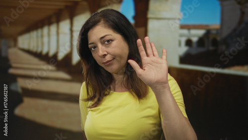 Middle aged hispanic woman waves hand at sunlit university colonnade in yellow shirt, standing along historic stone walkway; greeting warmth. Free Download Middle aged hispanic woman waves hand at sunlit university colonnade in yellow shirt, standing along historic stone walkway; greeting warmth. - GFXLoad