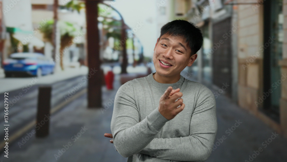 Fototapeta premium Young chinese man standing on a city street with buildings in the background, wearing a gray sweater and crossing his arms, showcasing a cheerful expression outdoors.