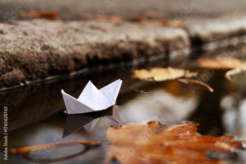 Paper Boat in a Puddle by a Curb with Autumn Leaves