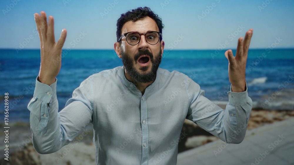 Naklejka premium Hispanic man with beard and glasses expresses surprise on a seaside promenade against a backdrop of blue sea and clear sky.