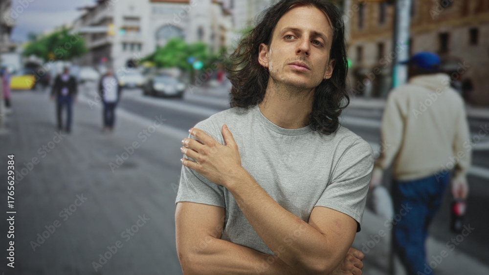 Fototapeta premium Man with long hair, hand to chin and crossed arms on city street, forearms exposed in resting pose; quiet contemplation.