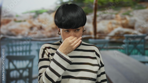 Photos Woman covering nose on outdoor restaurant terrace with striped sweater, expressing displeasure at bad smell, emphasizing the discomfort in a casual dining setting