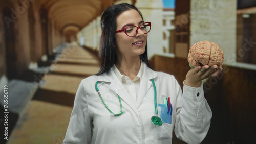 Hispanic woman doctor in uniform holding brain model with stethoscope in old university outdoor setting.