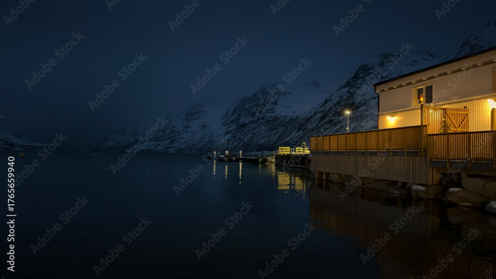 Fototapeta premium A winter night fjord in Norway. The mountain slopes are covered with snow. A small building is illuminated at night.