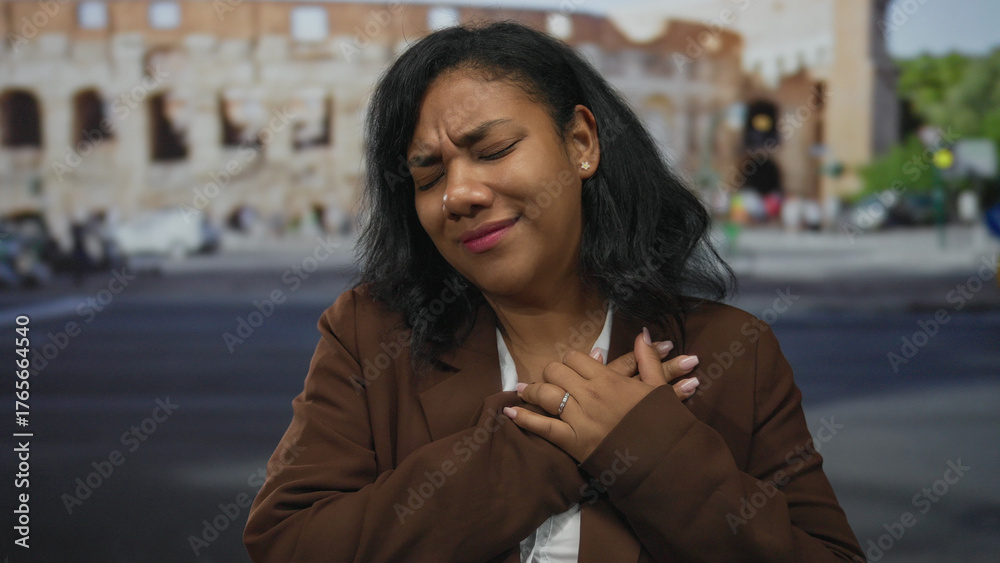 Fototapeta premium Woman clutching her chest with closed eyes and furrowed brow in front of roman coliseum outdoors during daylight; pain.