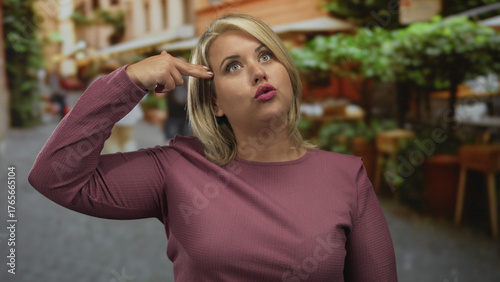 Fotografie Young blonde woman finger pointing to head on crowded street with terrace cafes; morbid despair suicide