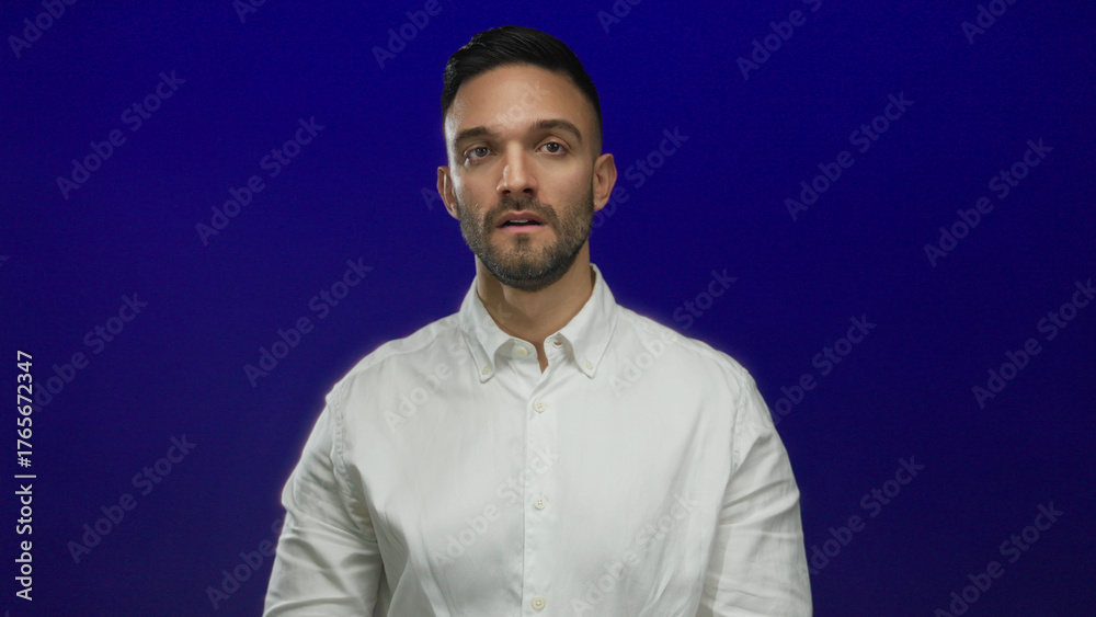 Obraz premium Tired young hispanic man in a white shirt poses thoughtfully against a solid blue background, conveying a sense of fatigue and introspection.