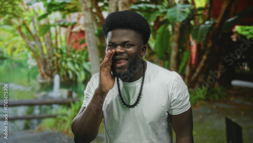 Man cupping hand to mouth shouting in forest park wearing white tshirt and beaded necklace; joy celebration.