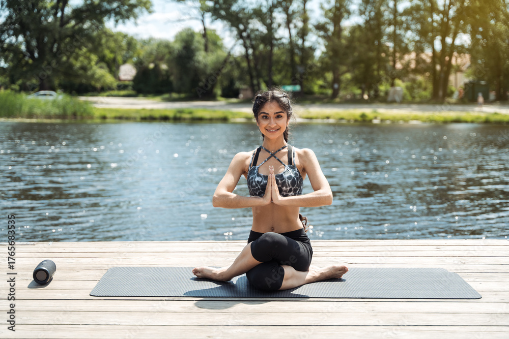 Fototapeta premium Young indian woman sitting in lotus pose meditating on wooden pier by the river. Outdoor yoga, relaxation, and mindfulness in nature.