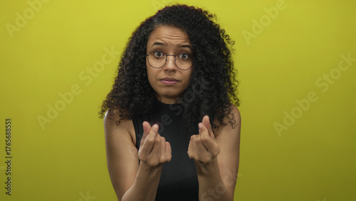 Fotografie Young hispanic woman rubs fingertips in studio over a yellow wall with worried glance; desperate plea for help