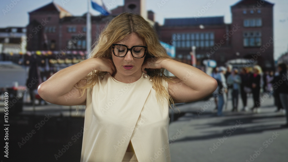 Fototapeta premium Woman adjusts neckline and touches neck wearing glasses on city street in front of brick building; contemplation.