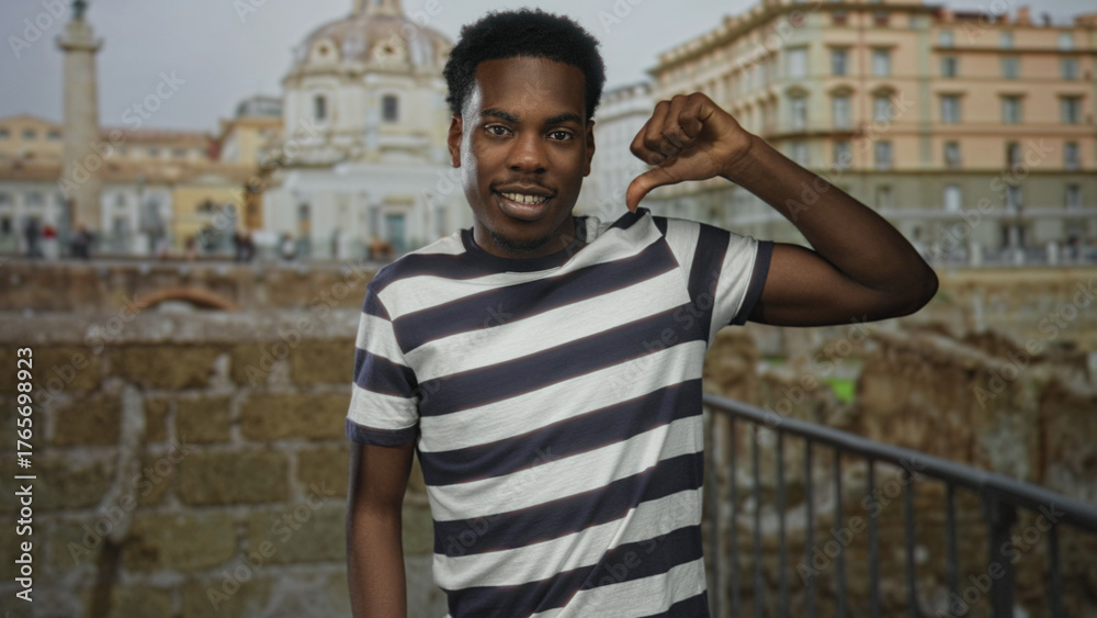 Fototapeta premium Man wearing striped shirt shows thumb down gesture at ancient building overlooking ruins; disapproval.